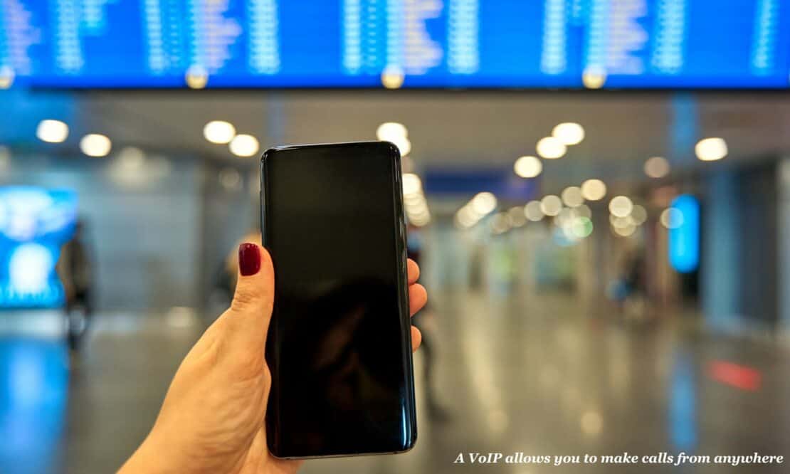 Woman holds a phone in her hand beneath an airport departures board - why a VoIP is essential for travellers