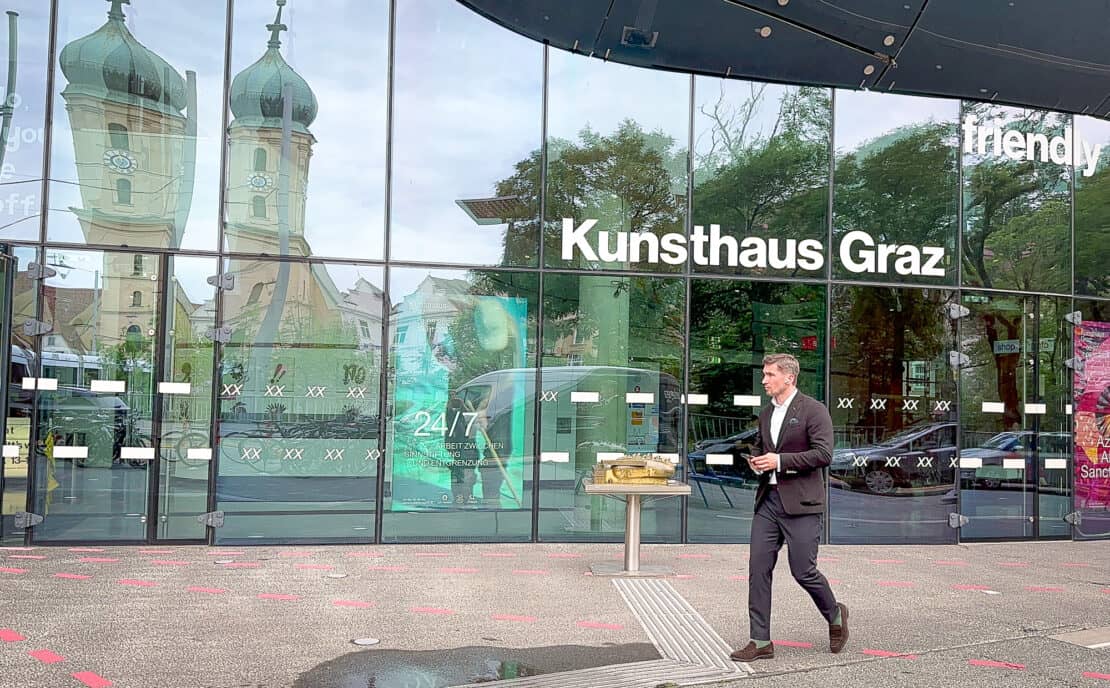 Man walking past Kunsthaus Graz with church reflection in glass